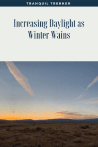 Desert scrubbrush and brown grass run to a silhouette of the mountain in the background. An orange sunset creates a backdrop, all under a clear, blue sky marked by jet contrails. Pin reads, "Increasing Daylight as Winter Wains."