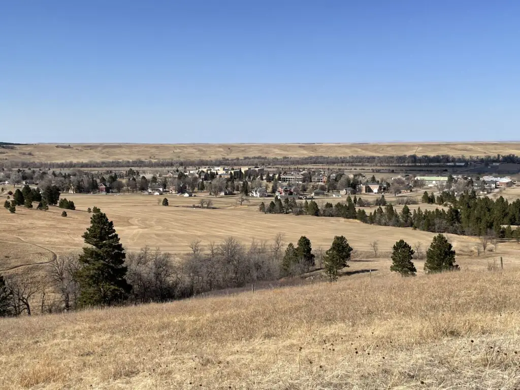 Brown-grass plain, green pines and leafless trees in the foreground, a small grouping of houses and other buildings in the background surrounded by trees