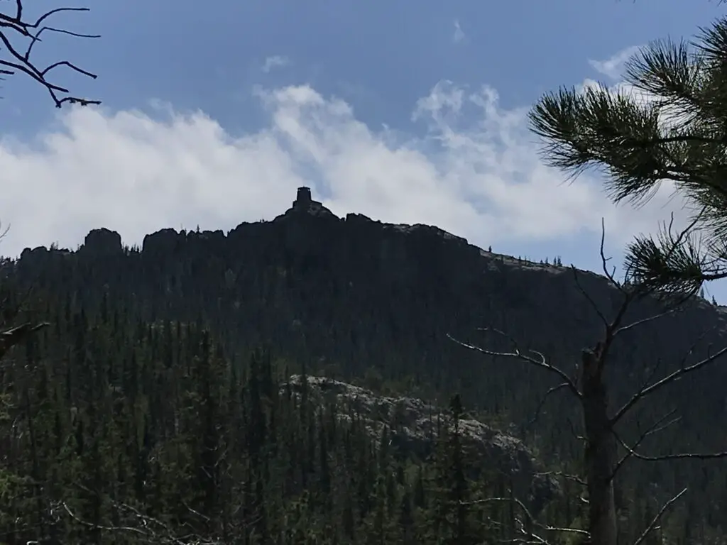 View from below: dark silhouette of a rocky, castle-like fire tower on top of a rocky, tree-covered mountain.