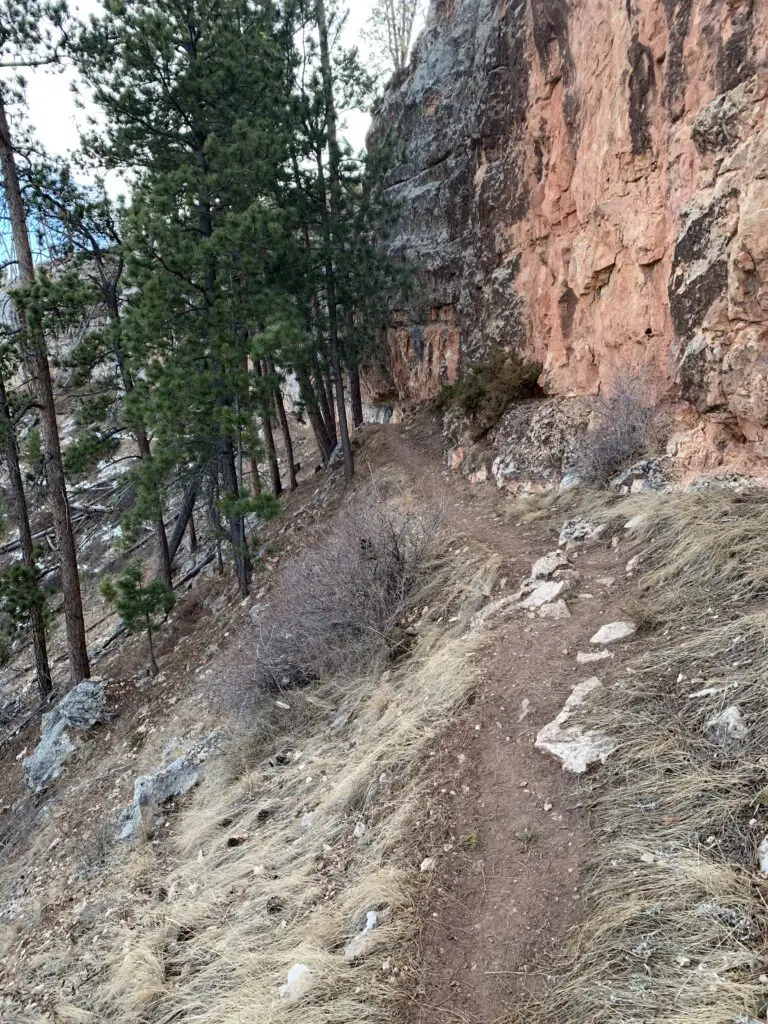 Dirt, walking path traverses the side of a steep, grassy hill. Red, rock walls rise on one side with pine trees on the other.