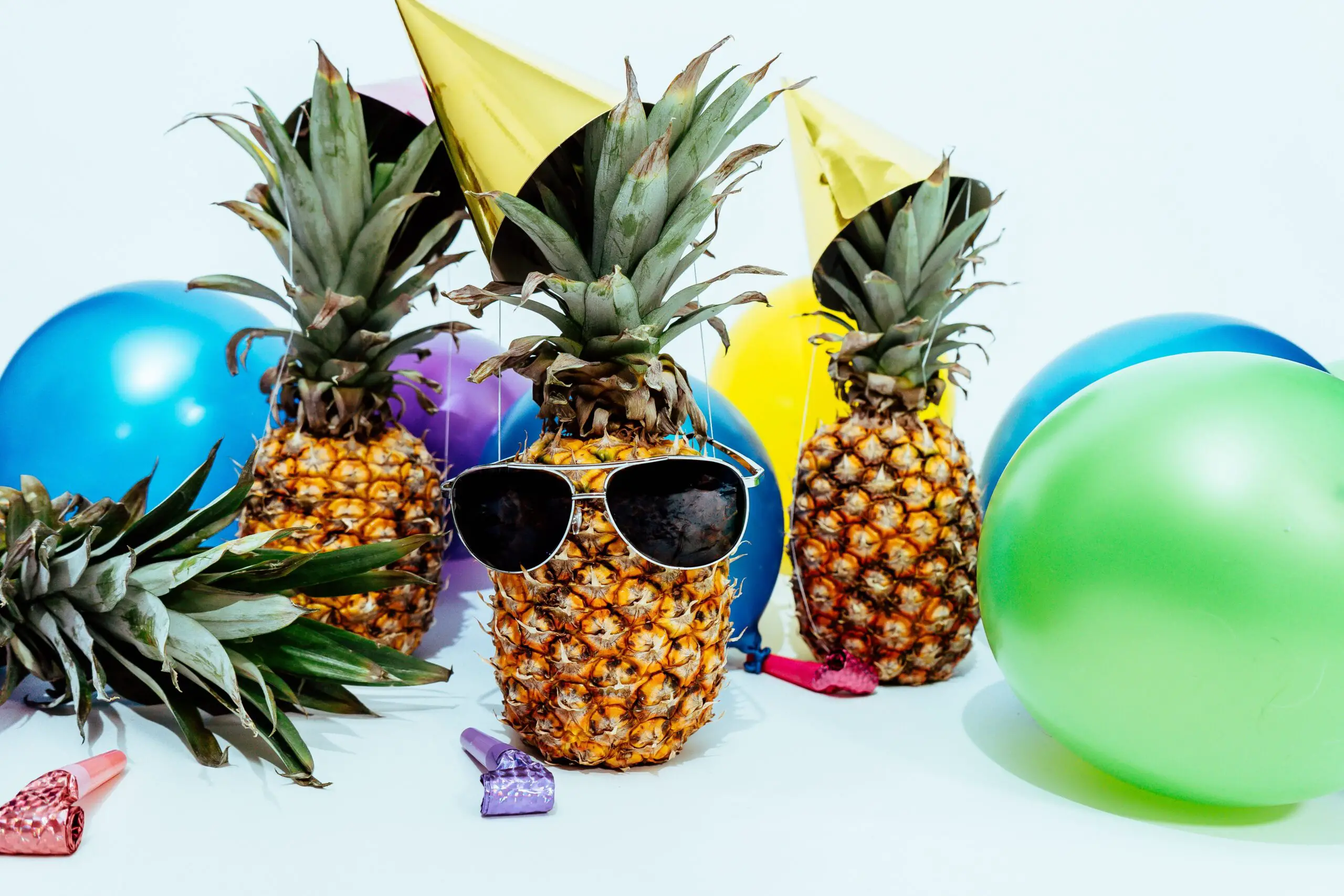 4 pineapples (one wearing sunglasses) are surrounded by balloons and party hats!