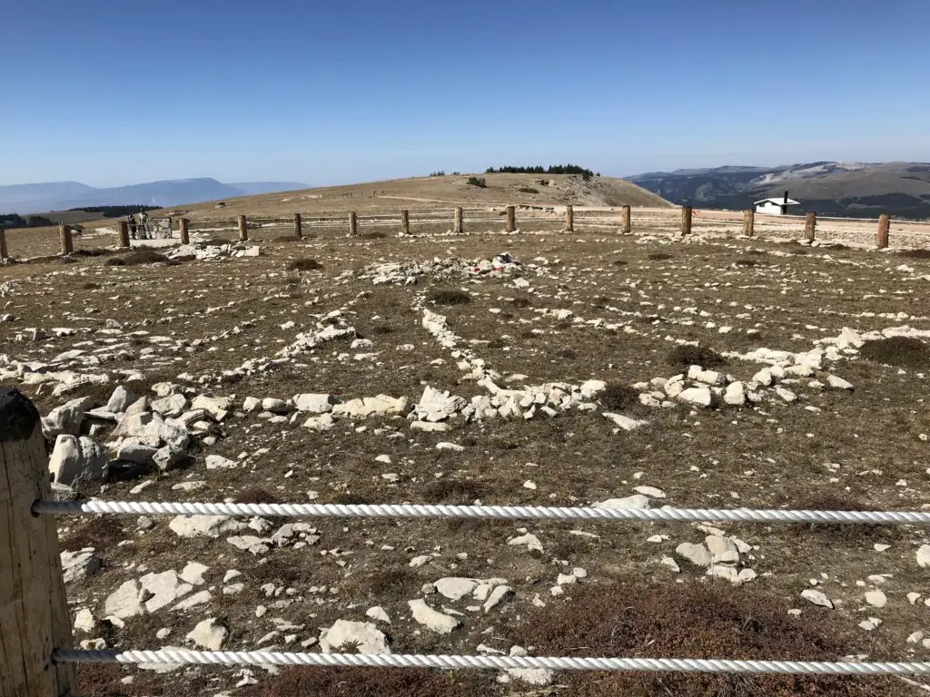 Small stones that form the shape of spokes on a wheel sit in a grassy area surrounded by a fence. Tall mountains are in the background, all sit under a clear, blue sky.