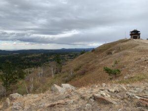 Dunn-colored rocks and dry grass in the foreground. A fire tower sits on top of the hill in the far backfound with pine tree-covered mountainsides.