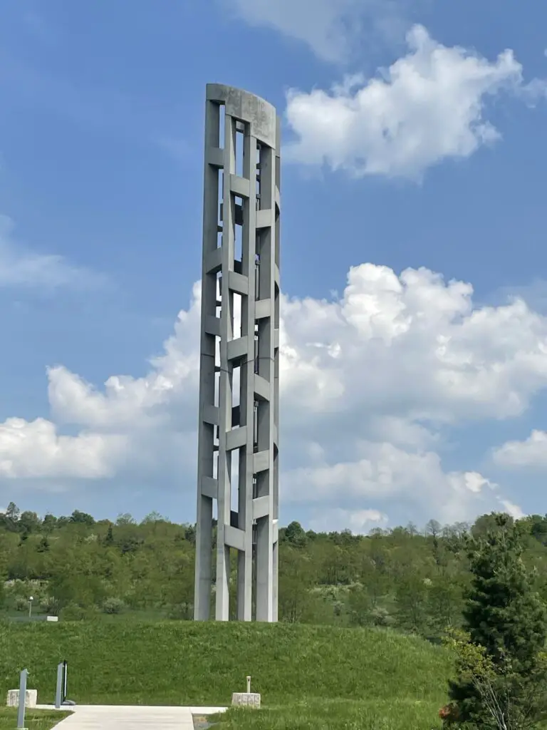 A tall, concrete tower with holes throughout and bells hanging from it sits on a grassy hill