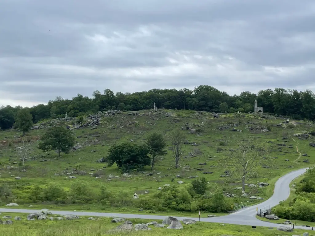 View from the bottom of a grassy hill that is speckled with large rocks and a few trees. More trees and stone monument line the top of the hill. 