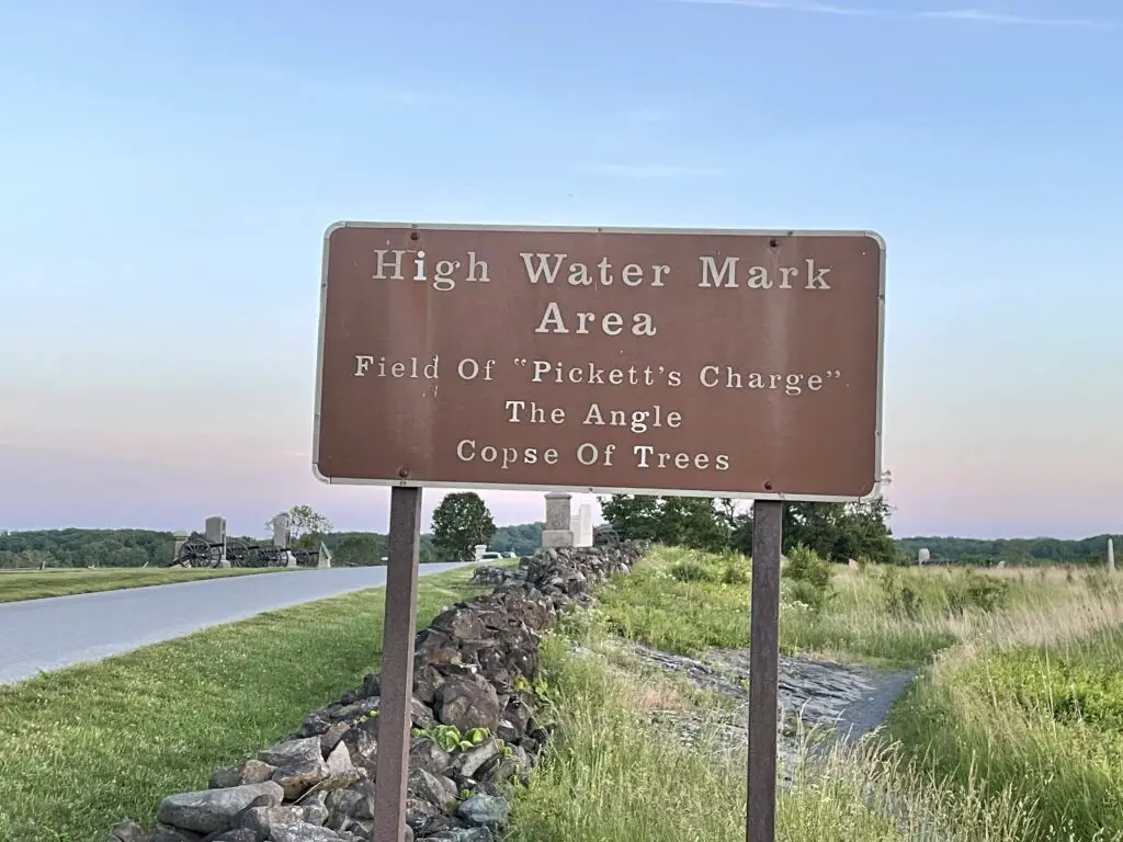 Brown informational sign in a field with a stone fence and stone monuments in the background. Sign reads, "High Water Mark Area. Fild of 'Pickett's Charge'. The Angle. Copse of Trees."