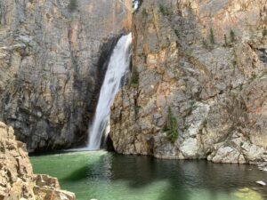 A thin waterfall pours through a split in some rocks into a greeninsh pool.