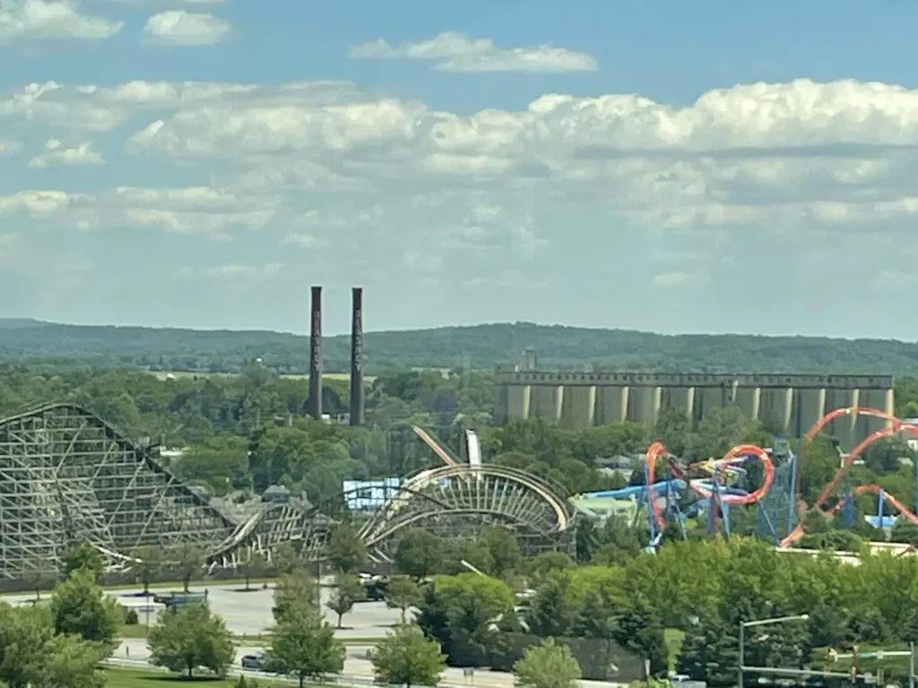 A view of multiple roller coasters amidst many trees. Green hills and smokestacks are in the background. 