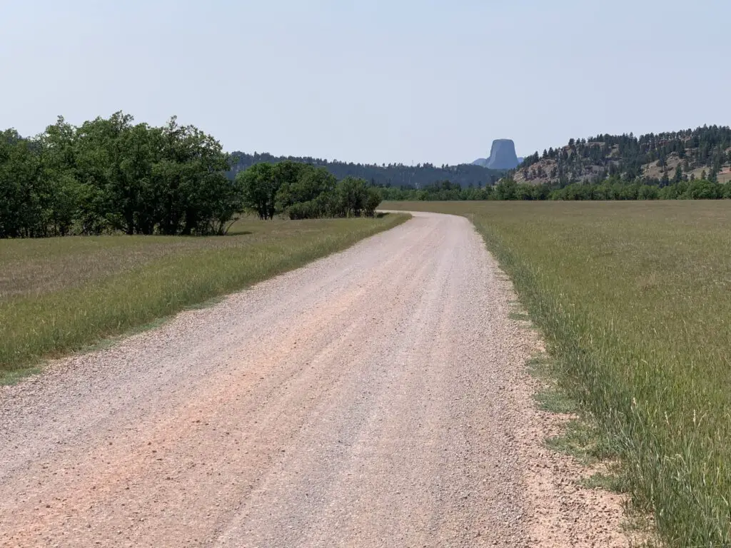 View down a dirt road that crosses a meadow. A rock obelisk (Devils Tower) materializes through the haze in the distance, behind tree-covered hills. 