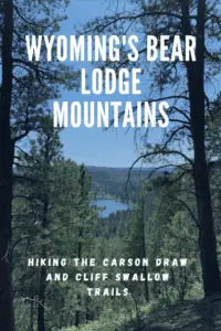 View down a tree-covered hill to a long, blue lake, surrounded by more trees, in the distance. Pin reads, "Wyoming's Bear Lodge Mountains Hiking the Carson Draw and Cliff Swallow Trails"