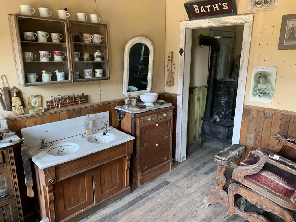 Interior of building. Sinks, mirror and wash basin in foreground, antique chair to the side, antique wood stove through doorway. Sign above door says, "Bath's"