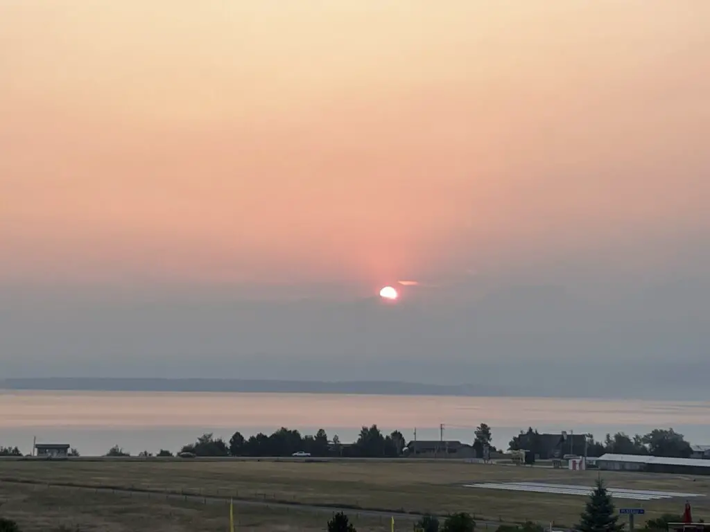 Grassy area with a large lake in the background. The sun rises over some shadows in the far background through the haze of wildfire smoke.