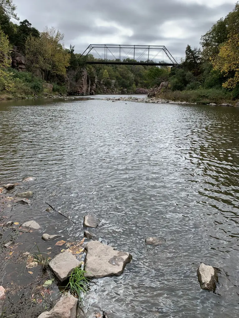 Metal bridge spanning creek, vegetation-covered rock walls on both sides; cloudy, grey sky.