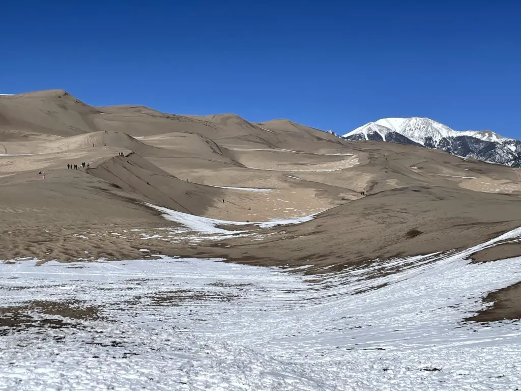 Snow-covered sand dunes with snow-covered mountains and a clear, blue sky in the background