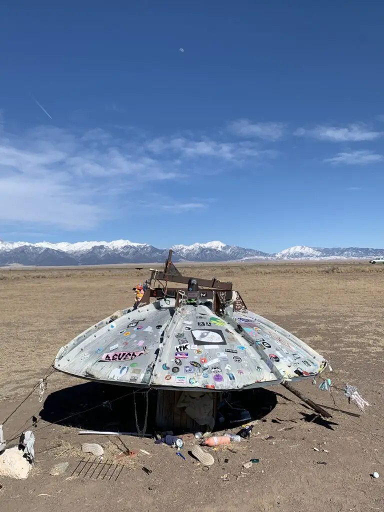 Old satellite dish covered with stickers and surrounded by trinkets in the foreground, snow-covered mountains far in the background