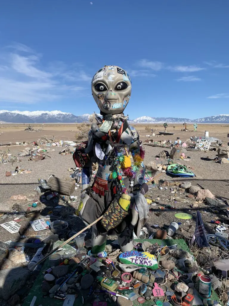 Alien figure surrounded by and covered in trinkets and figurines