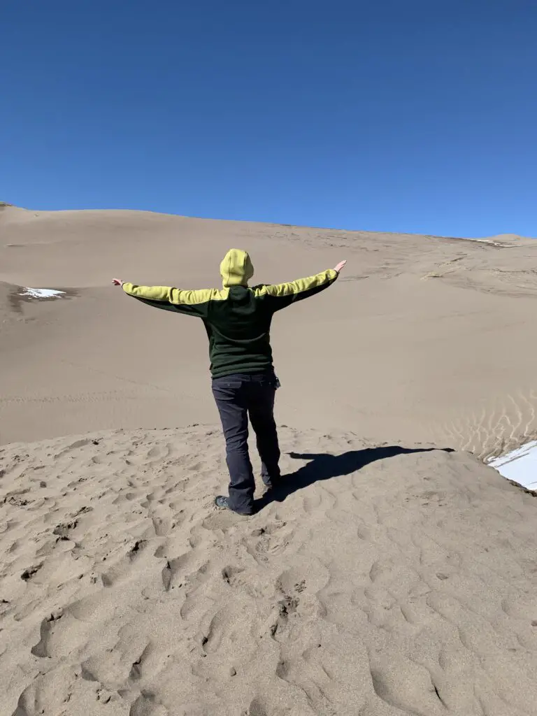 Person with back to camera standing with arms-outstretched atop a sand dune.