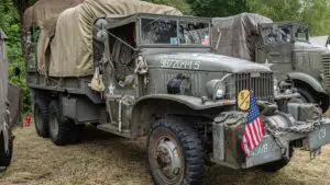 An army truck with a small, American flag on the front sits near other army trucks in a woodlike setting.