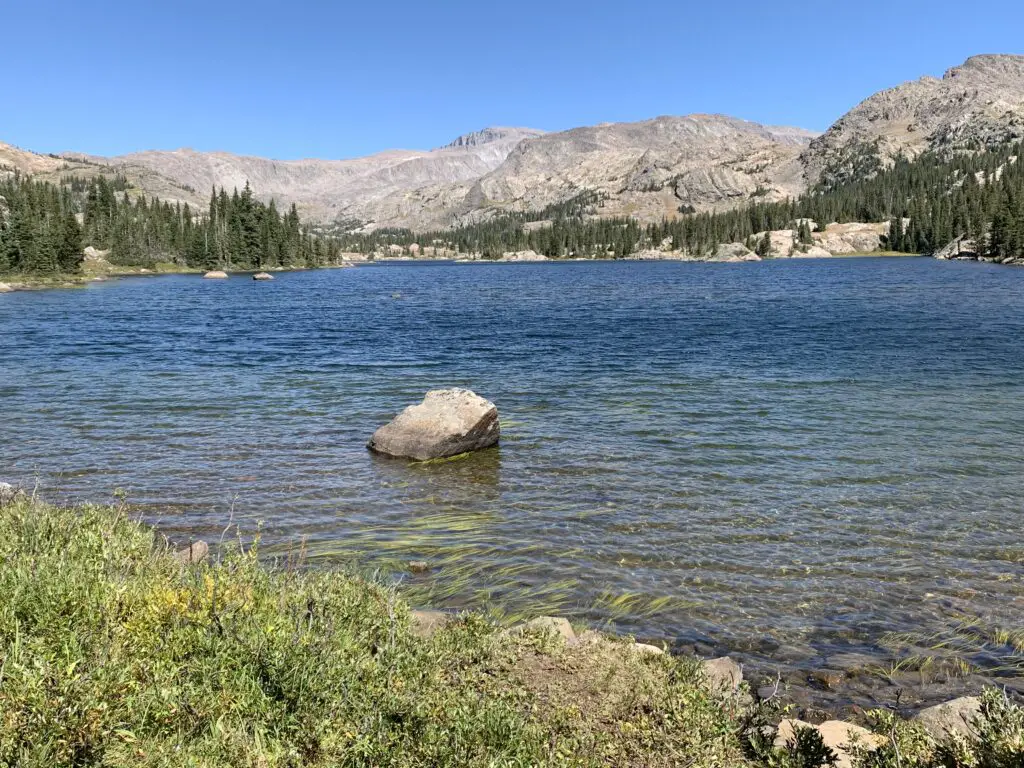 Small lake with a large rock in the middle, ringed by trees. Rocky mountains rise in the background.