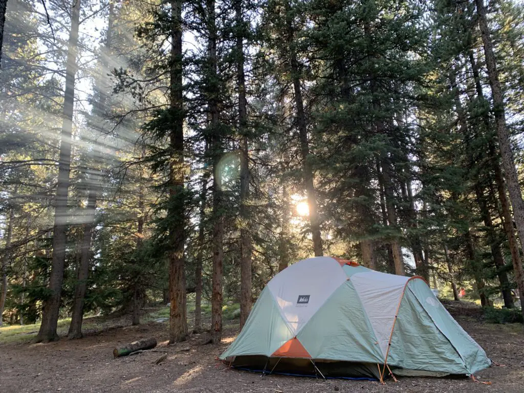 A tent sits in the forest, sunlight streams through the trees and creates beams in the smoky air