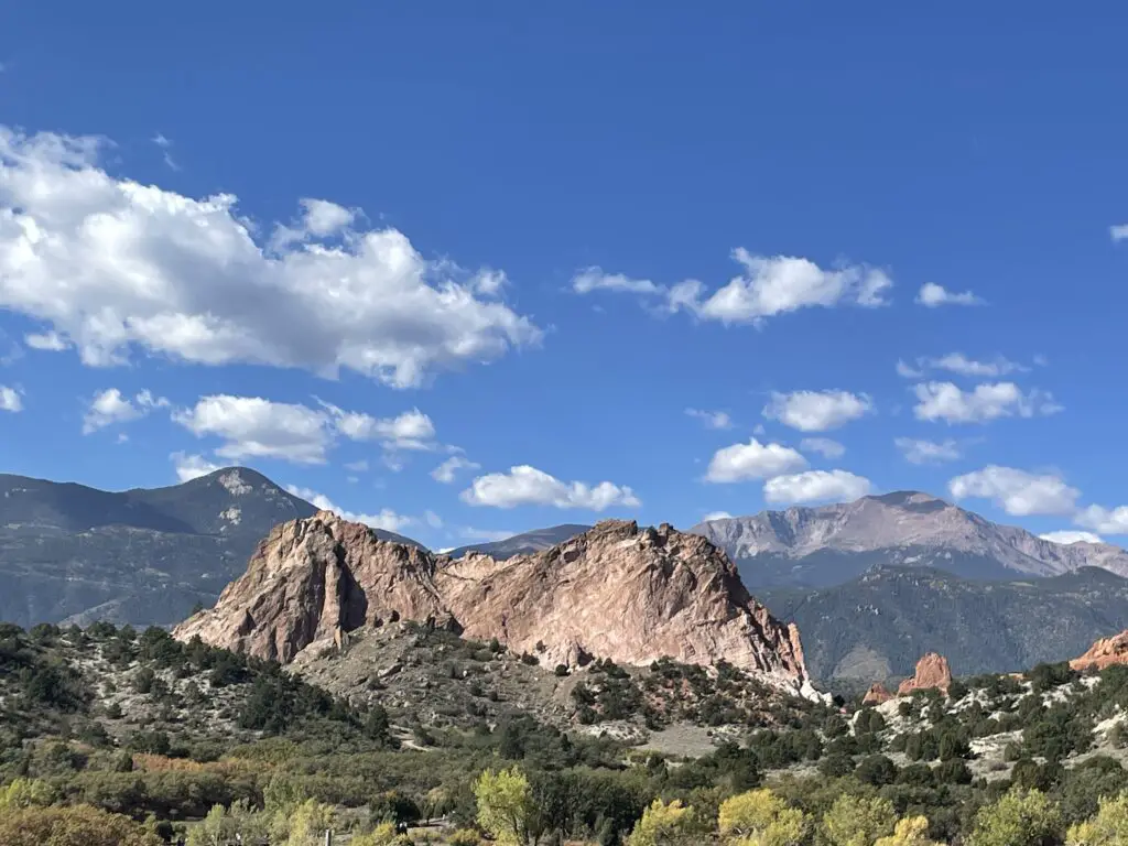 Green, scrub brush with a large rock rising out of it in the foreground, much taller mountains in the background including one that rises above tree line. 