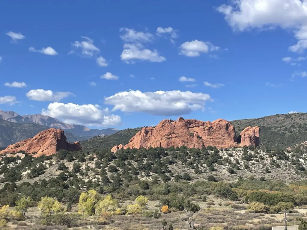 Red rock shapes rise above green scrubland. Two shapes that resemble camels with their heads touching sit on the top of one of the red rocks.