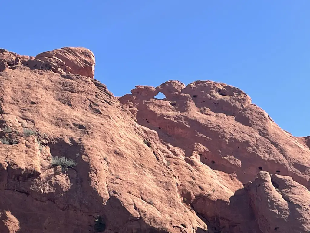 Red rock wall. Two shapes on top resemble the head of a cow and a bird with its wings back sit facing each other with their heads touching.