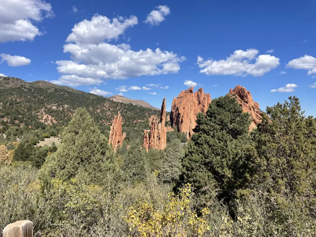 Red rocks rise from green, scrub brush. A tree-covered mountain in the background has a taller, rocky mountain rising behind it.