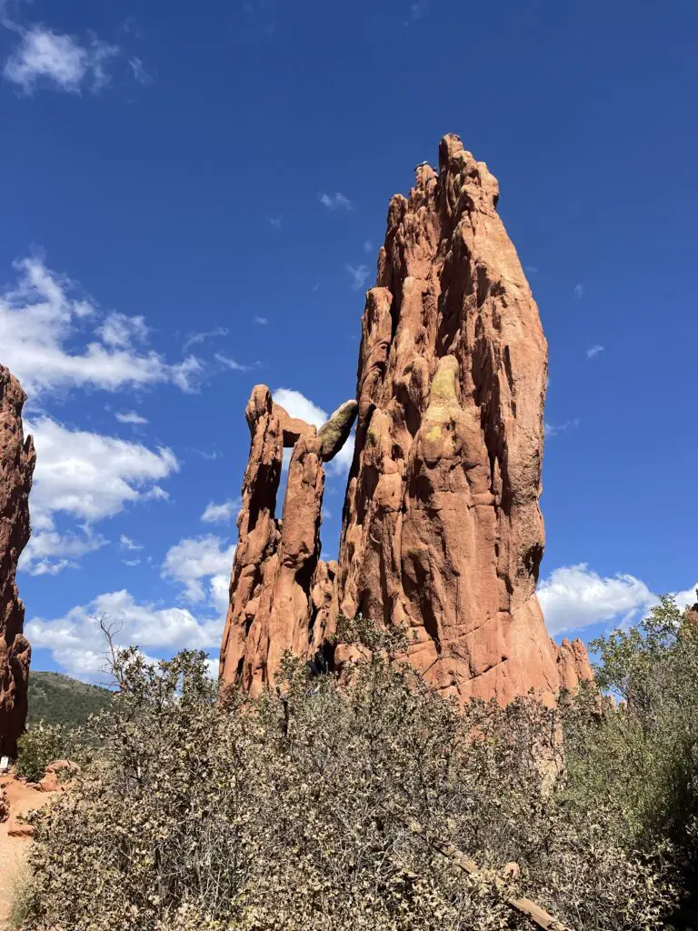 Tall, narrow, red rocks rise from a scrub brush, desert landscape on a blue sky, white, puffy cloud day. One rock has a hole through the middle. 
