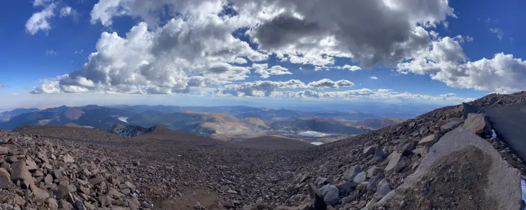 Panoramic view of rocky slopes of a mountain in the foreground. Lower mountains and plains are in the background with puffy, white clouds and blue sky overhead.