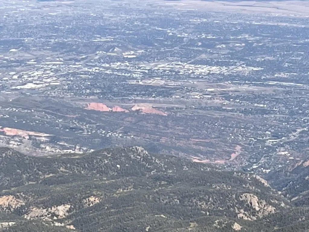 Hazy view of tree and grass-covered hills. A city and red rocks rising from the green are in the distance and far below. 