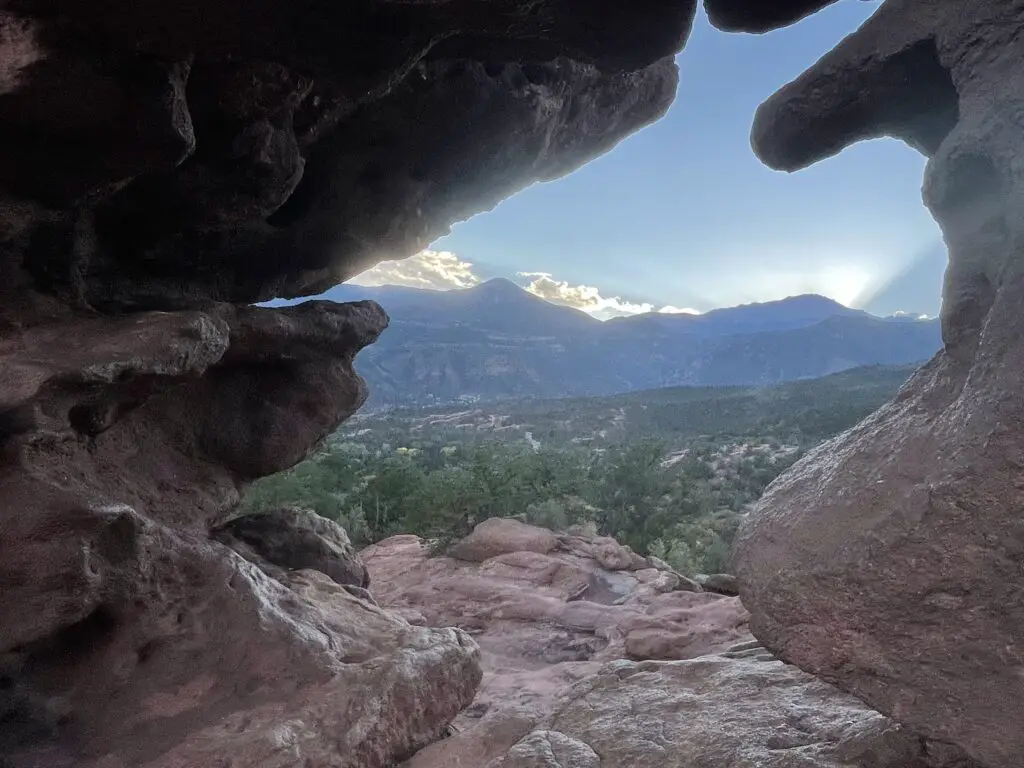 Dark, mountain silhouettes viewed through a whole between two tall, narrow rock formations. The mountains in the background tower over the green, desert landscape in the foreground.