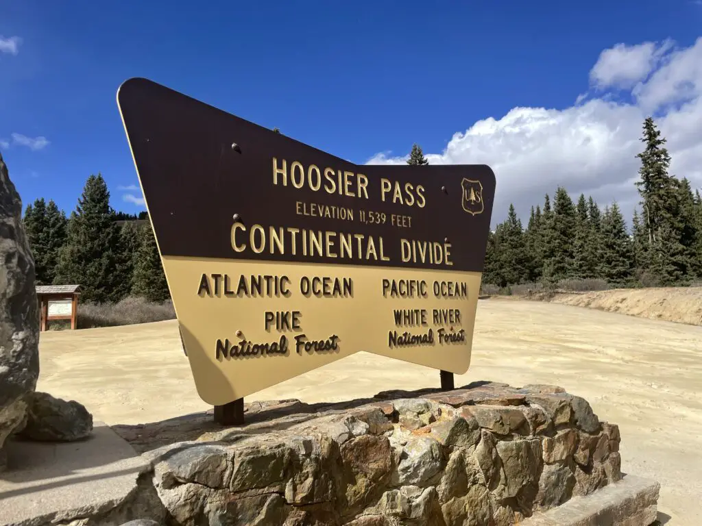 Informational sign on a rock at a gravel parking area reads, "Hoosier Pass elevation 11,539 feet. Continental Divide Left: Atlantic Ocean Pike National Forest Right: Pacific Ocean White River National Forest
