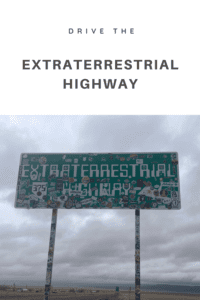 Green highway sign covered in stickers. Can barely make out the words "Extraterrestrial Highway". Pin reads, "Drive the Extraterrestrial Highway"