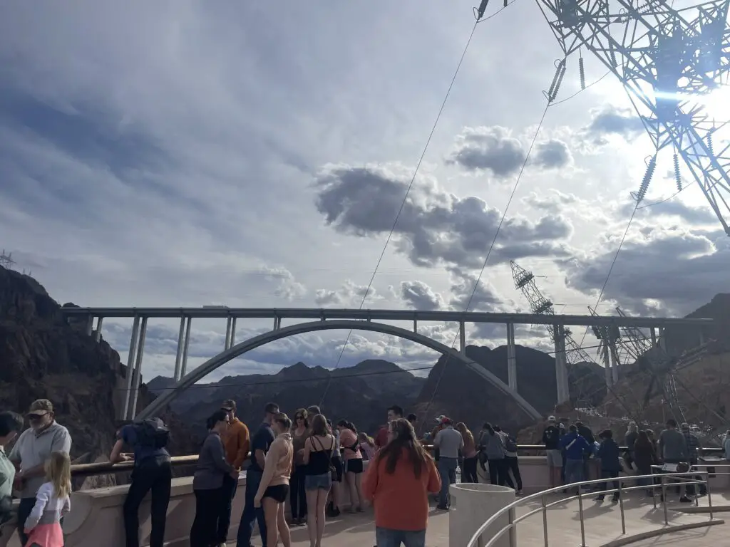 A bridge spans a rocky canyon. People stand on an observation deck in the foreground with high-tension electrical towers and wires rising overhead