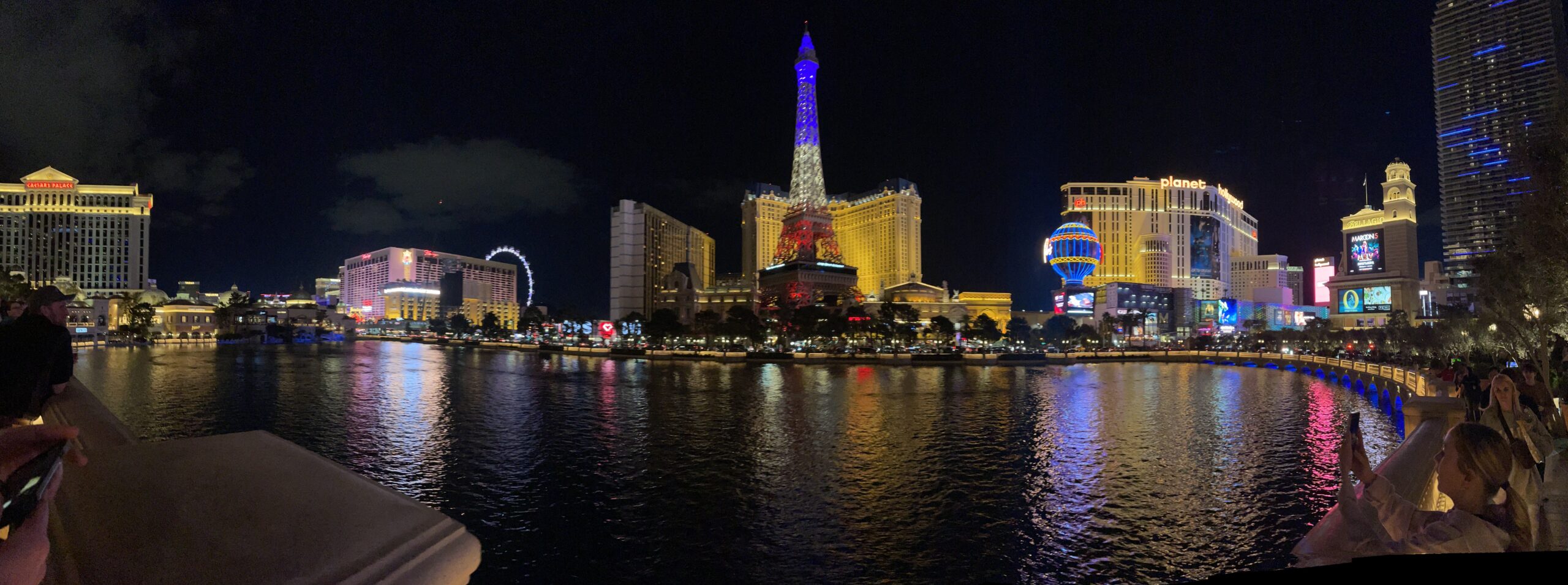 The neon lights of the Vegas Strip surround a pool of water and are reflected in it, at night. 
