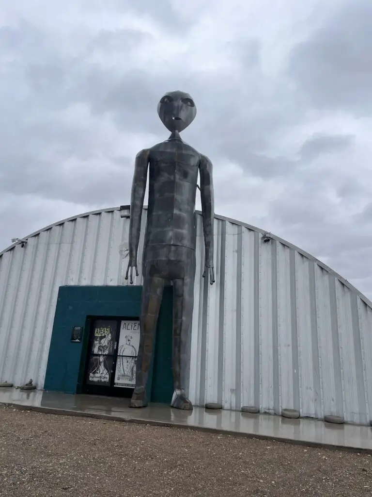 Two-story tall, metal alien stands outside a small building. 