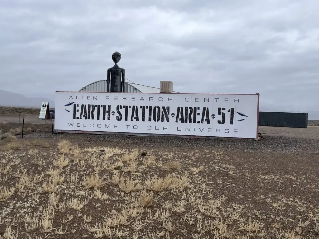 Sign in the foreground reads, "Alien Research Center, Earth Station Area 51, Welcome to our Universe". The top of a 2-story tall, metal alien stands behind the sign.