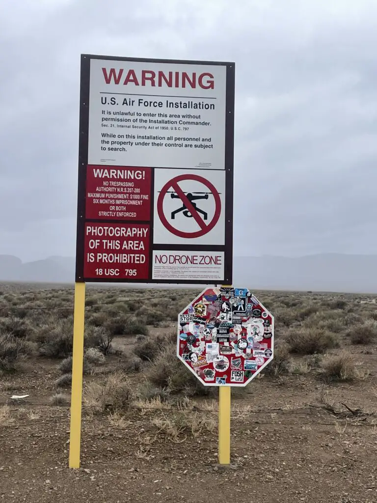 A stop sign covered in so many stickers it is almost unreadable. Another sign above it reads, "WARNING, US Air Force Installation. PHOTOGRAPHY OF THIS AREA IS PROHIBITED. NO DRONE ZONE.