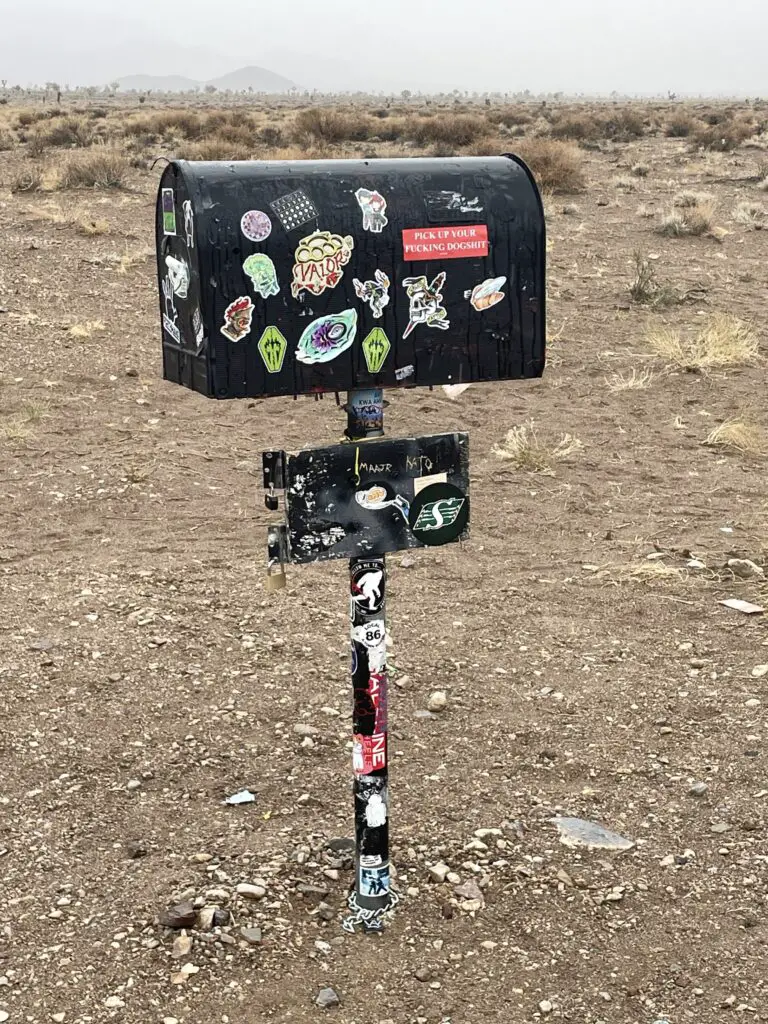 A black mailbox covered in stickers sits in the desert