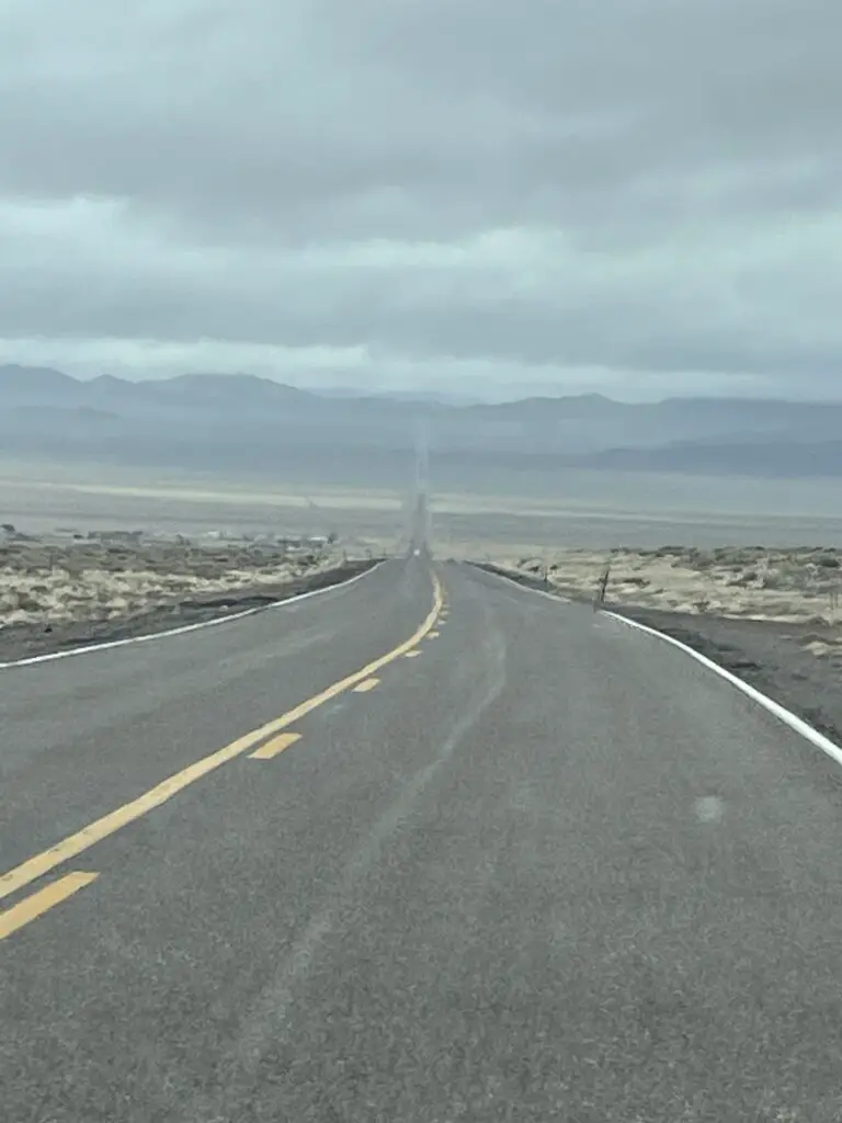 An empty, two-lane road stretches across the desert, to the horizon and up a hill towards mountains in the distance. 