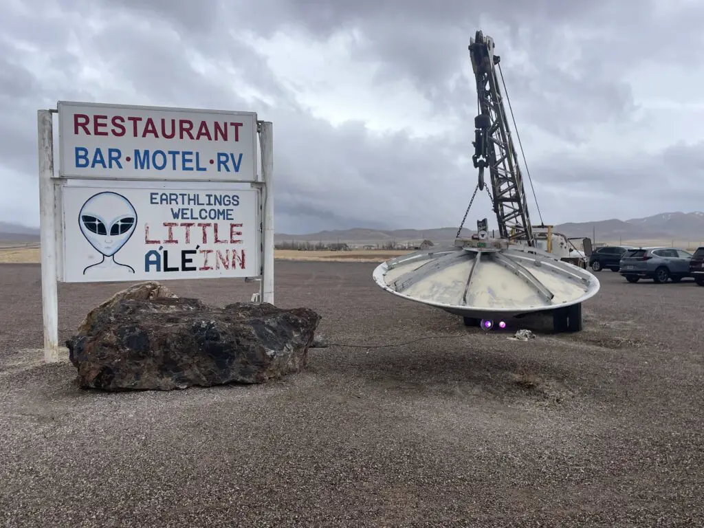 Parking lot with a sign that reads, "Restaurant, Bar, Motel, RV." A drawing of an alien and the sign reads, "Earthlings Welcome. Little AléInn". Also, a very old, upside down satellite dish hangs from a small crane, appearing like a small flying saucer.