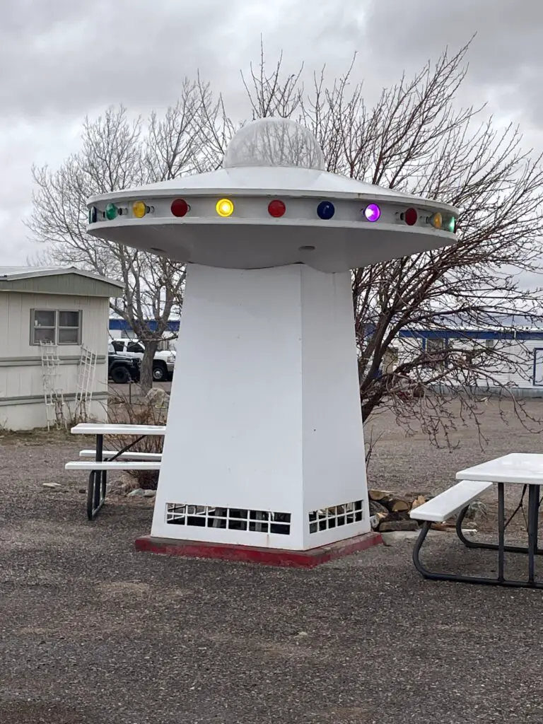A small flying saucer with lights sits on a short pedastal near some picnic tables