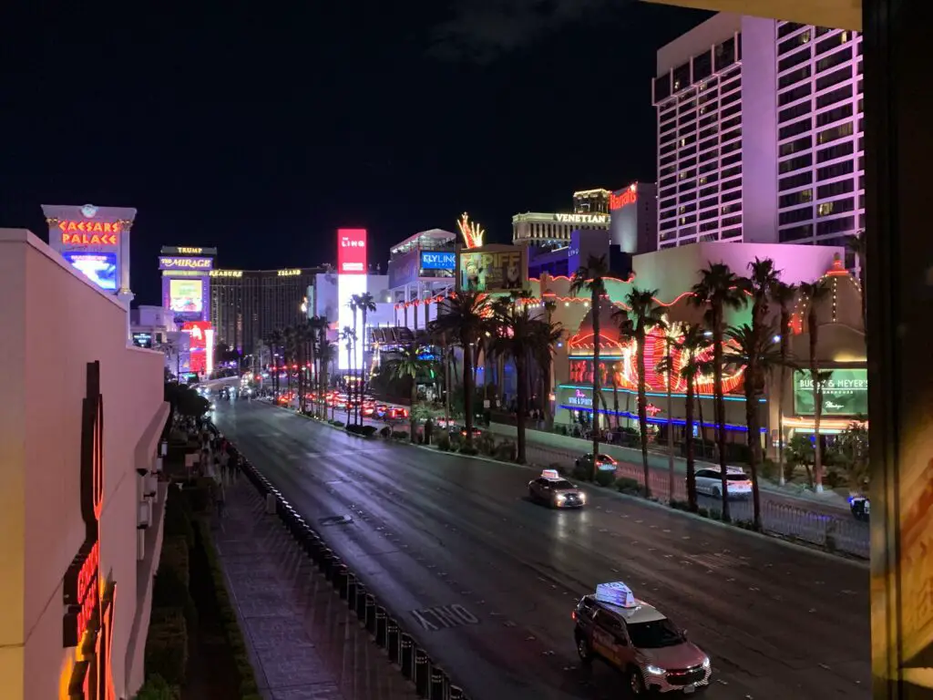 A large roadway with a row of palm trees in the middle and surrounded by the neon-lit, Vegas Strip, at night. 