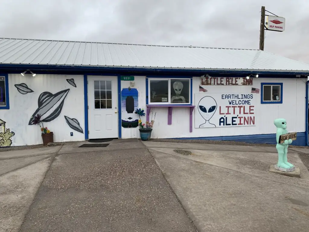 A small restaurant with alien and flying saucer drawings, A short, green alien statue stands in front and a sign reads, "Little AléInn, Earthlings Welcome". 