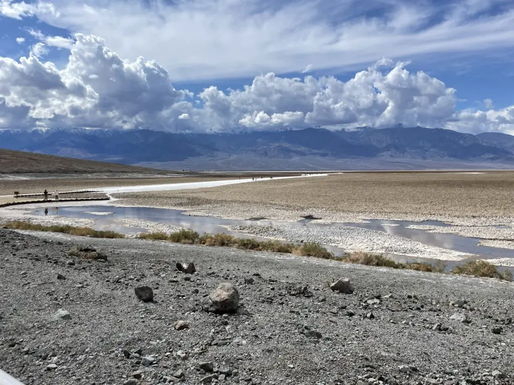 A rocky, salt-flat spreads to the rocky mountains on the horizon. A puddle of water reflects the sunlight in the foreground.