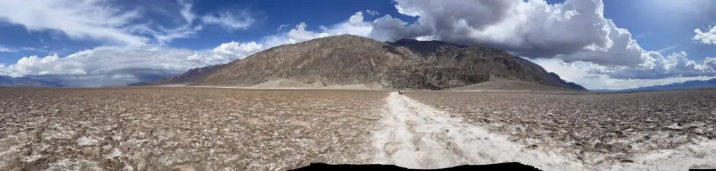 Panoramic picture of a flat valley, filled with a salt-flat, and sand. A salt-flat path heads toward a towering, rocky mountain.