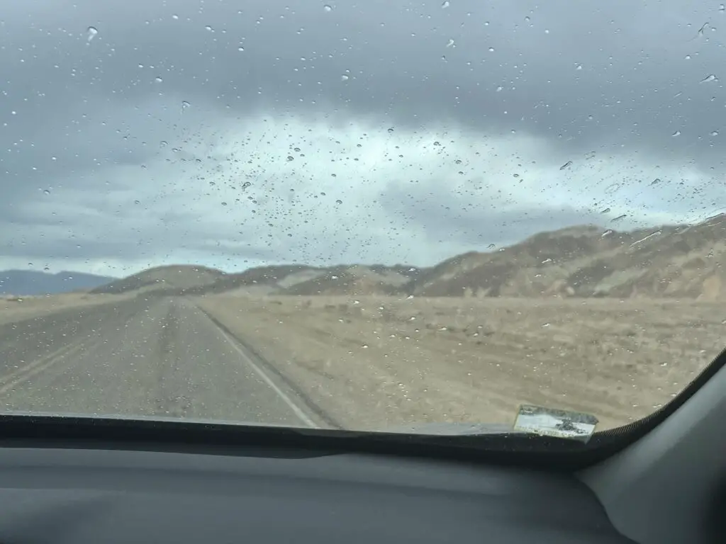 View through the front window of a car onto a road, a brown, sandy desert, and hills. Raindrops spatter the windshield