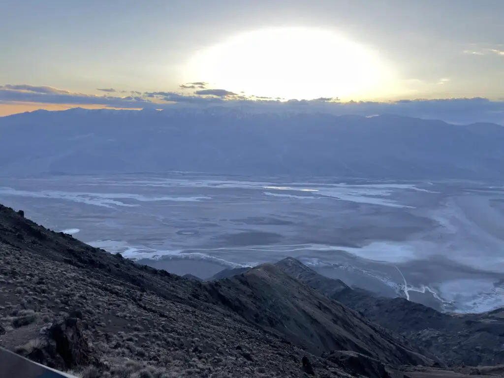 Grey valley streaked with salt stains leads to dark, shadowed mountains. A yellow, sun arc rises out of these mountains and clouds as the sun sets.