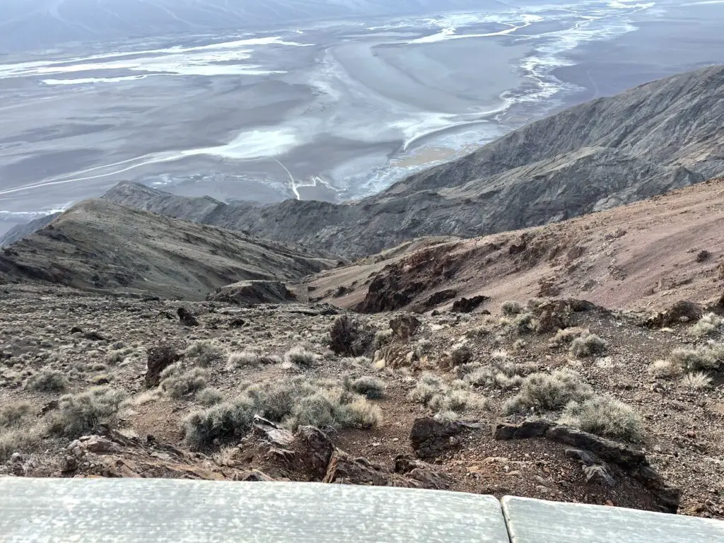 View from above, looks down a rocky and scrub brush-covered cliff to a gravely plain streaked with salt stains and water puddles